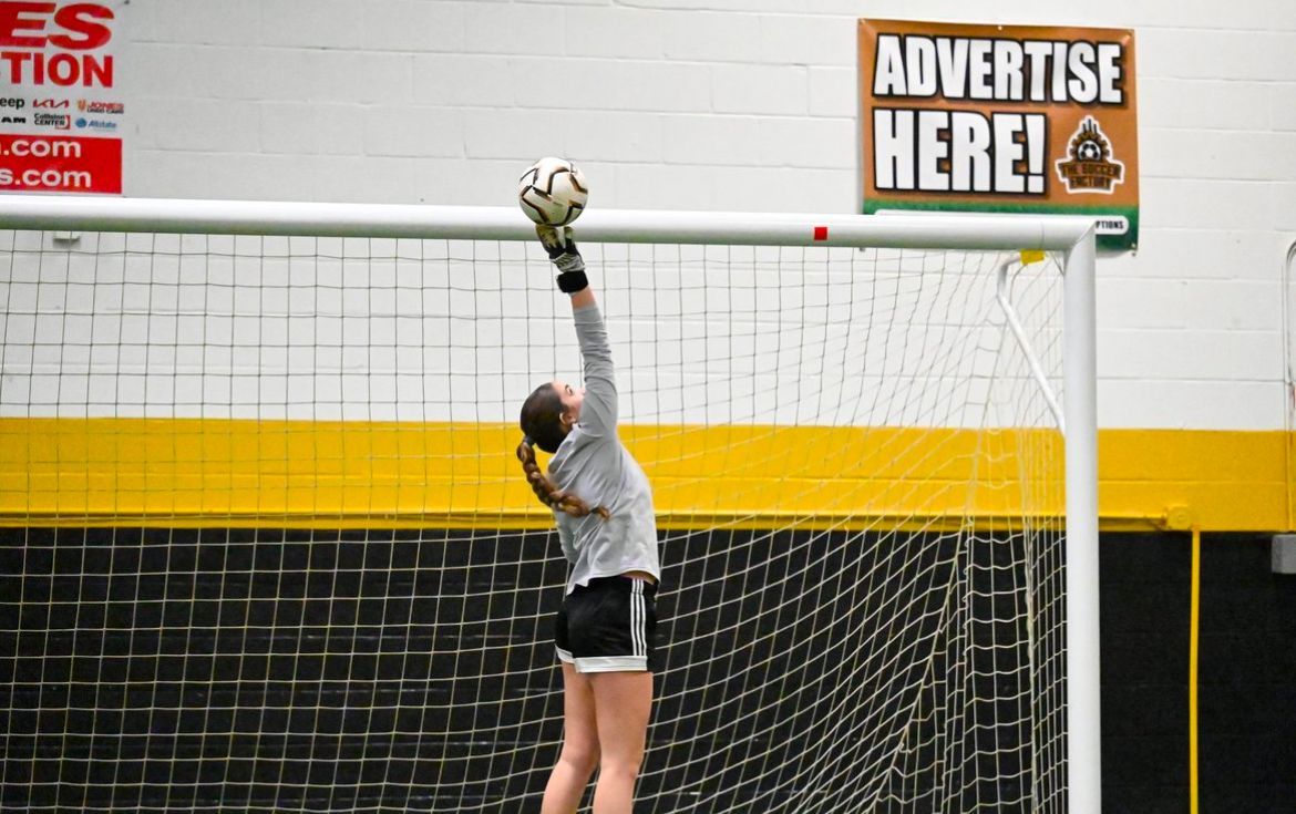 A soccer goalie catches a ball in front of a sign that says advertise here