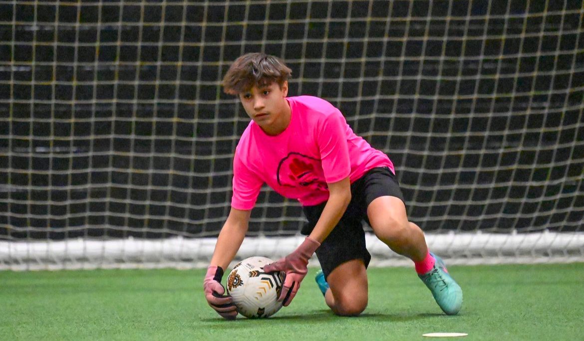 A young boy in a pink shirt is kneeling down to catch a soccer ball.