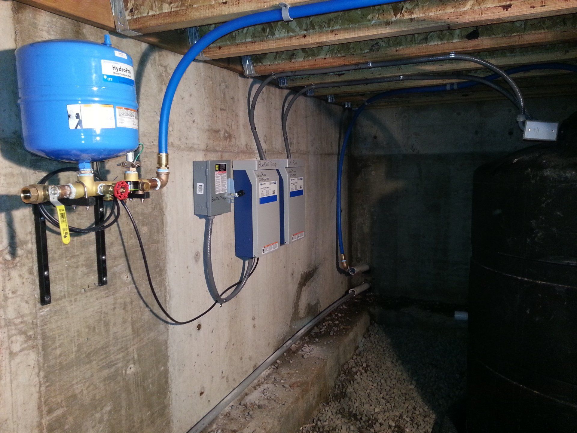 Well water system in a basement, including a blue pressure tank, electrical control boxes, and plumbing. The setting is concrete and dimly lit.