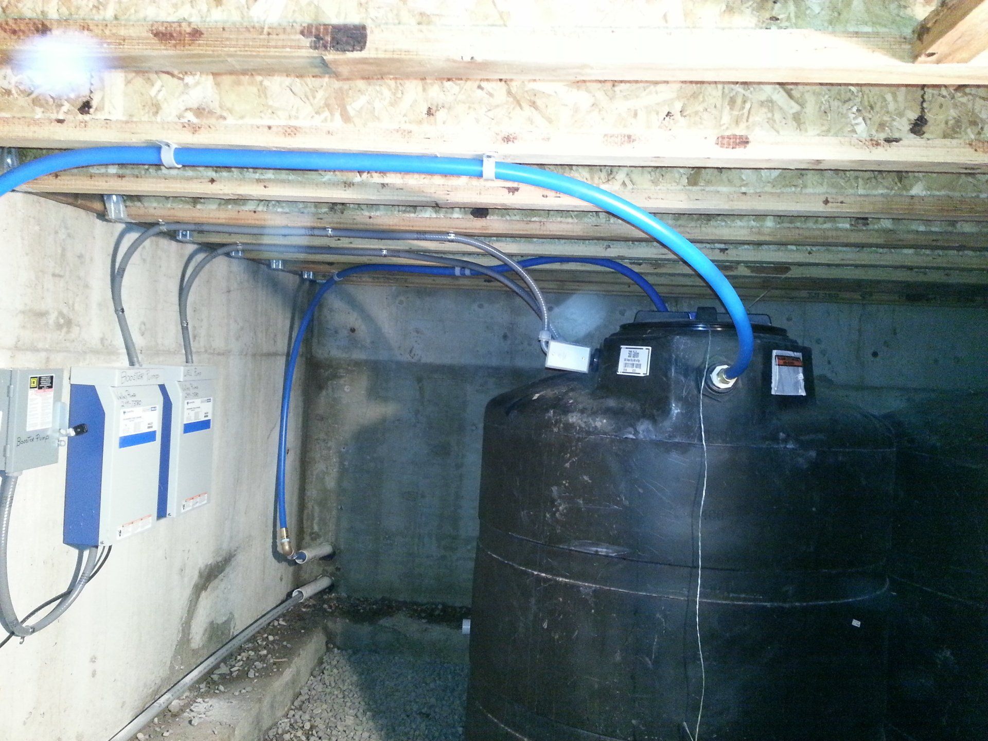 Black water storage tank in a basement setting, connected to blue pipes and electrical components on the wall.