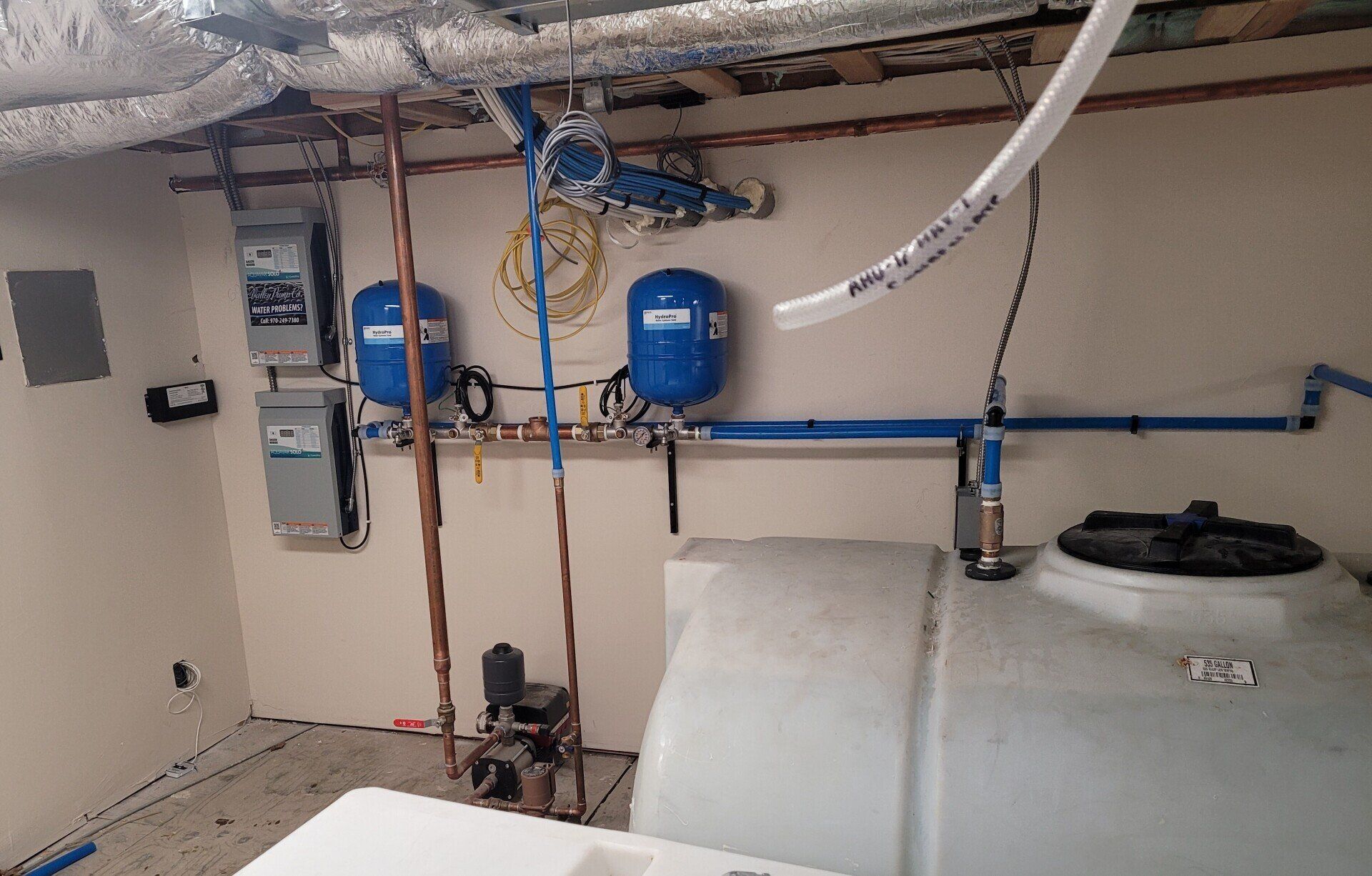 Plumbing equipment in a utility room: water tanks, pressure tanks, and electrical boxes against a white wall.