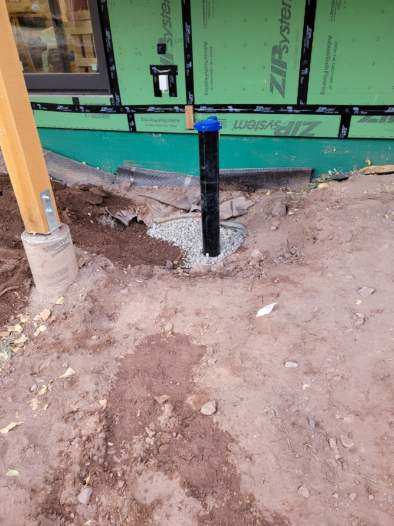 Black pipe with a blue cap emerging from gravel, in a dirt yard with a wooden post. Green building material is in the background.