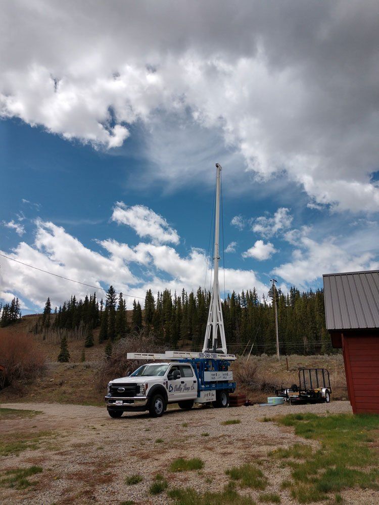 A white well drilling rig on a truck is set up in a rural area with green trees and a cloudy blue sky.