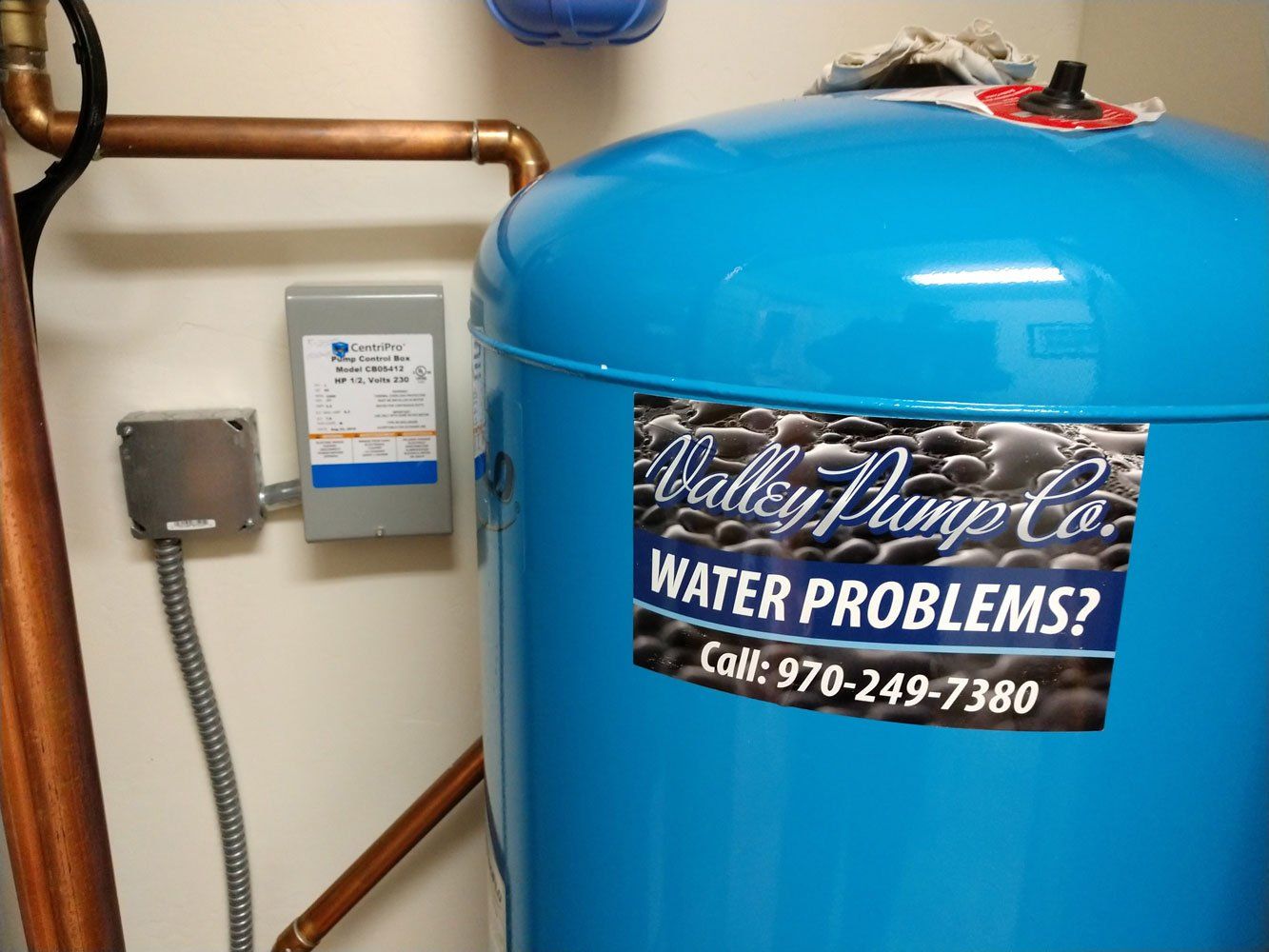 A blue water tank with a Valley Pump Co. sticker next to a wall-mounted control box and copper pipes.