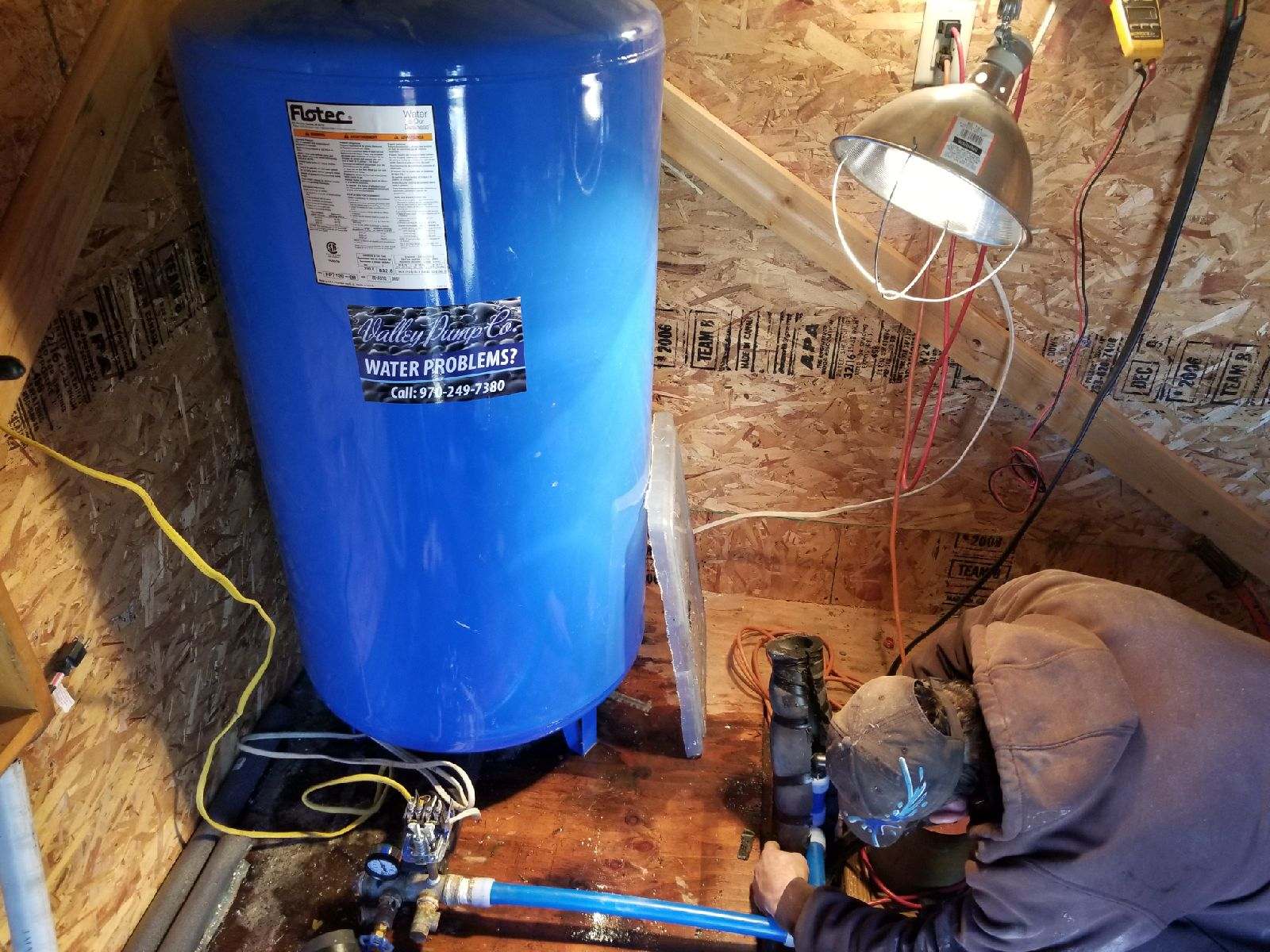 A person in a hooded sweatshirt works on plumbing near a blue water tank in a dimly lit utility room.