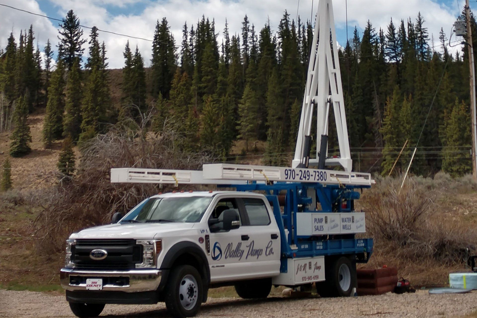 A white well-drilling truck with a tall drilling tower in front of a forest. The truck has blue and white detailing.