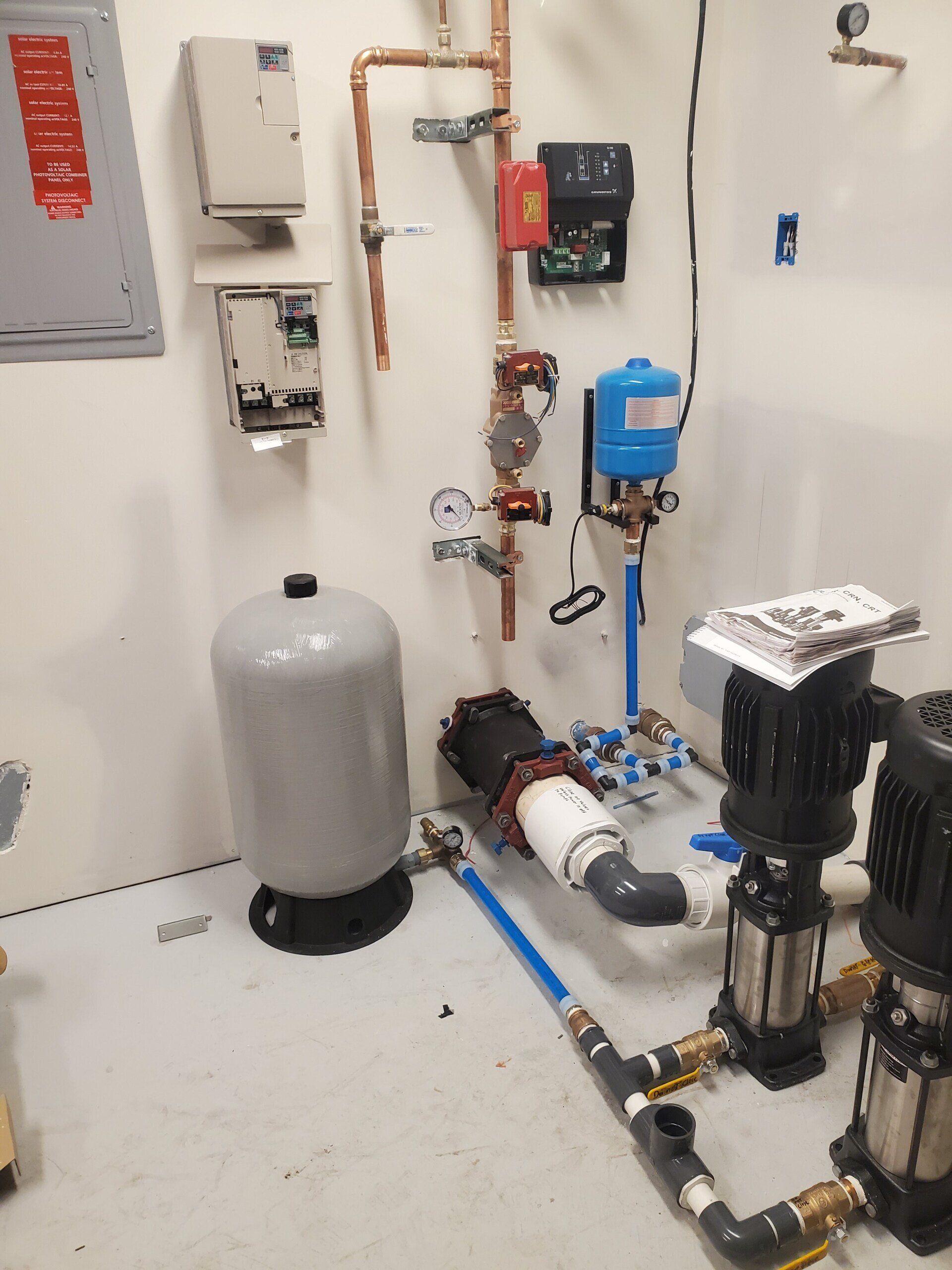 Plumbing system with water tanks, copper pipes, pumps, and electrical components against a white wall in a utility room.