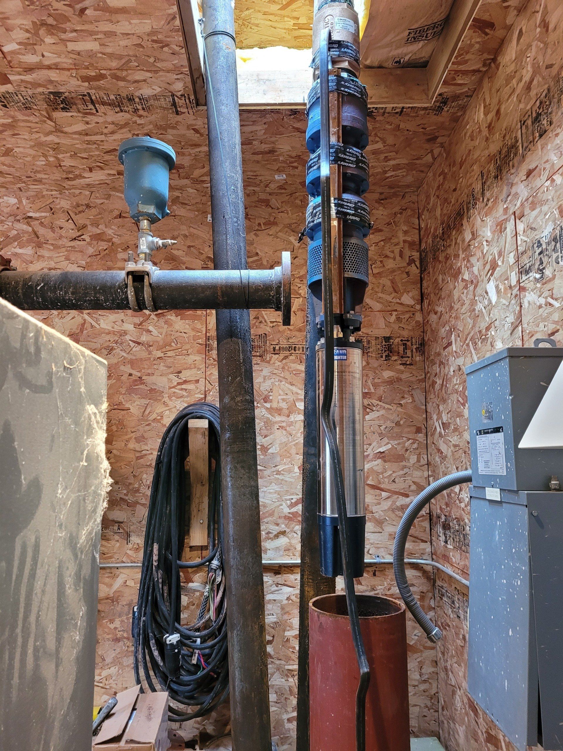 A water pump system in a wood-paneled room, with pipes, wires, and a blue pressure gauge.
