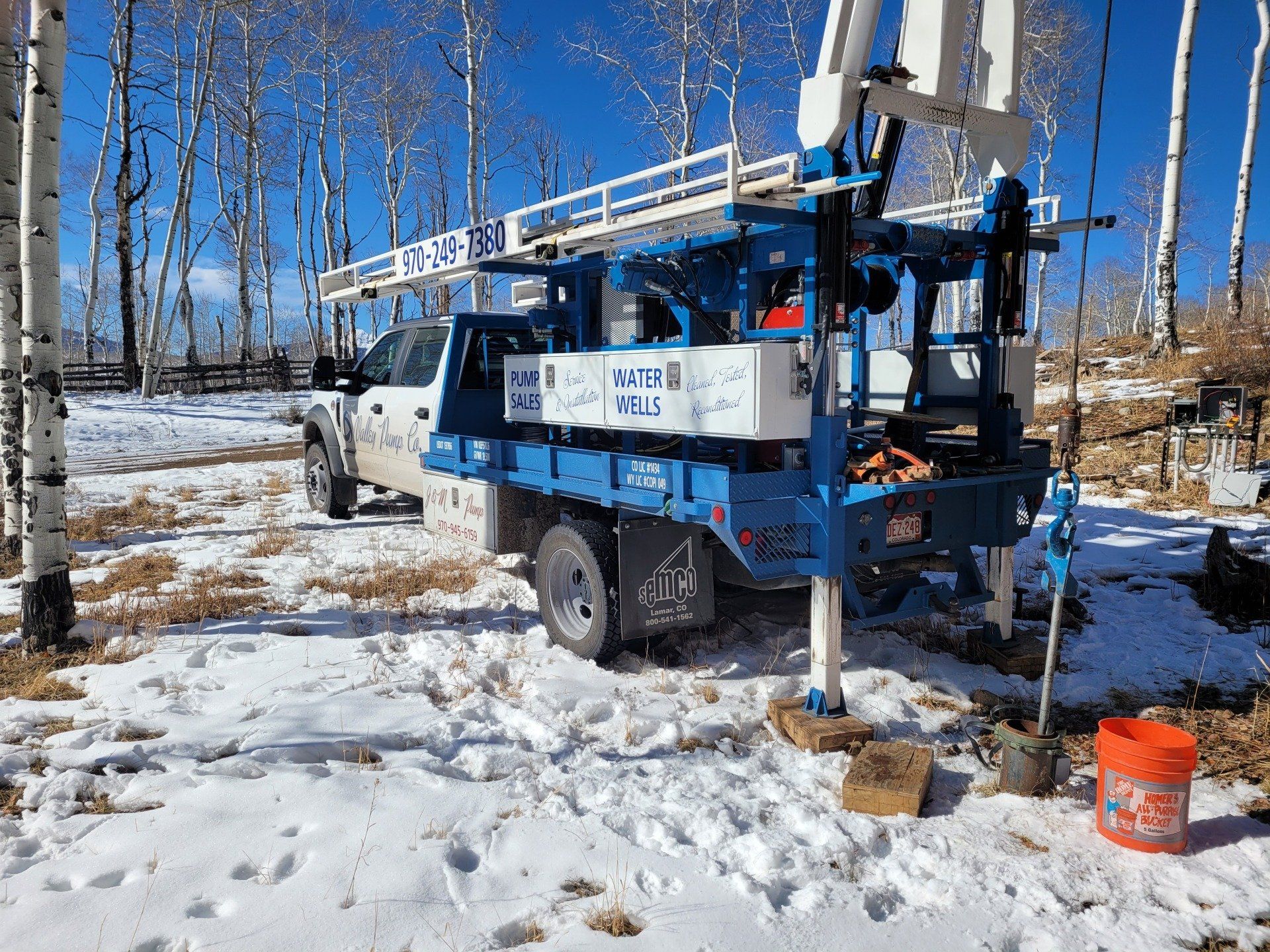 Blue and white drilling rig truck parked in a snowy forest with aspen trees. An orange bucket sits nearby.