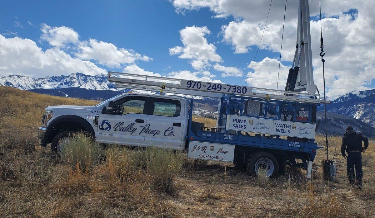 White and blue service truck with ladder on a hillside, drilling rig extended. Mountains in the background, person standing next to rig.