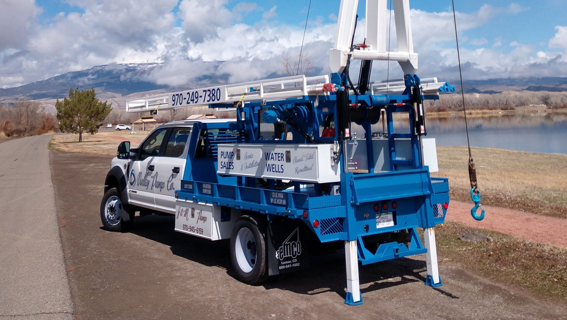 A white and blue drilling rig mounted on a truck parked on a paved road next to a lake and mountains.