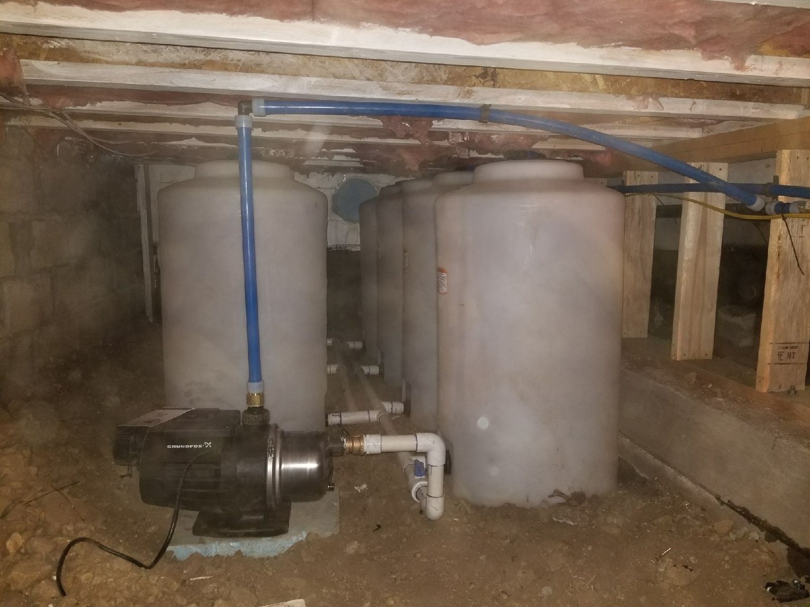 Four white water storage tanks connected by blue pipes and a water pump in a crawl space.