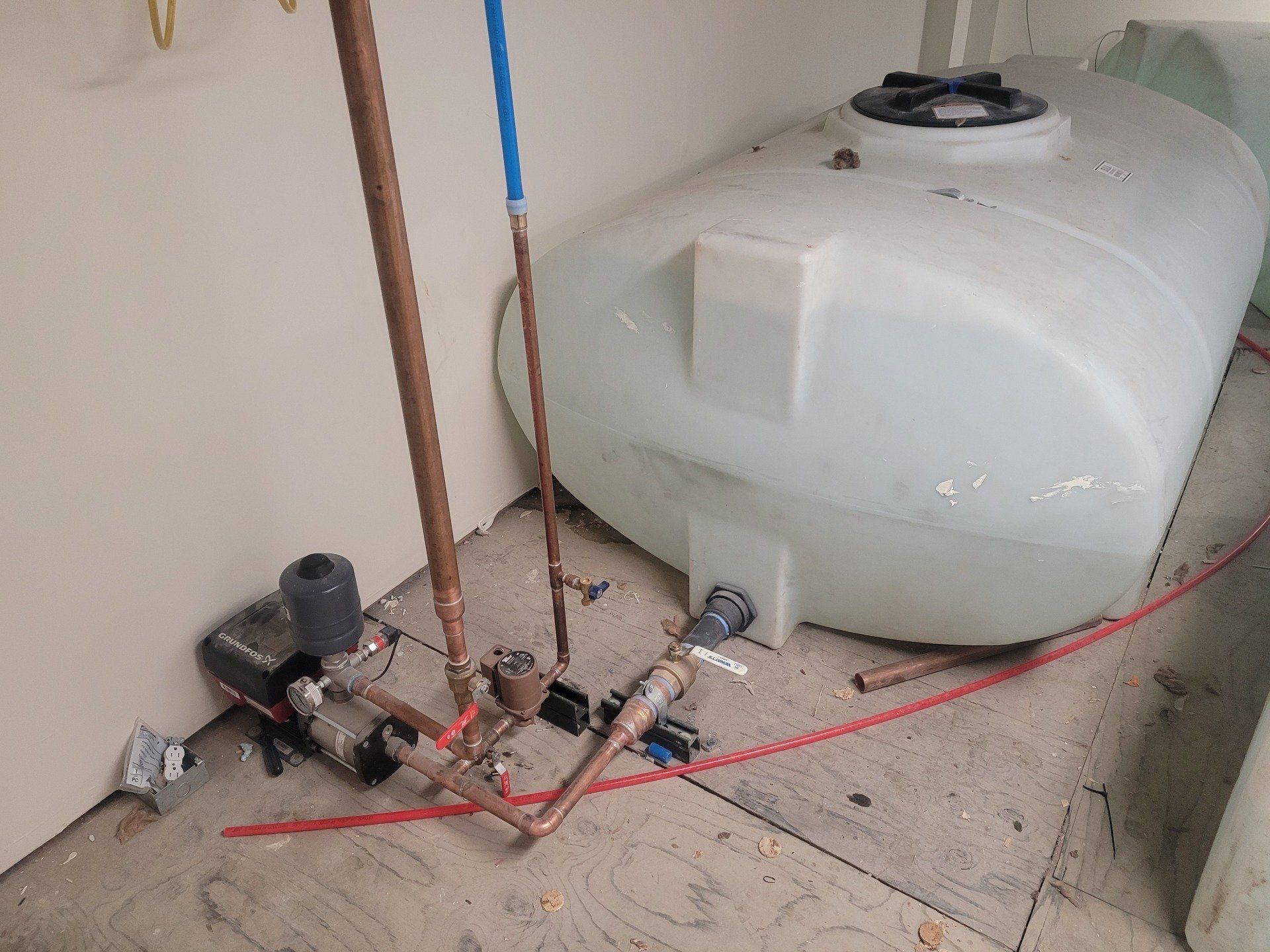 White water tank with associated plumbing in a corner of a room. Copper pipes, blue tubing, and a pump are visible.