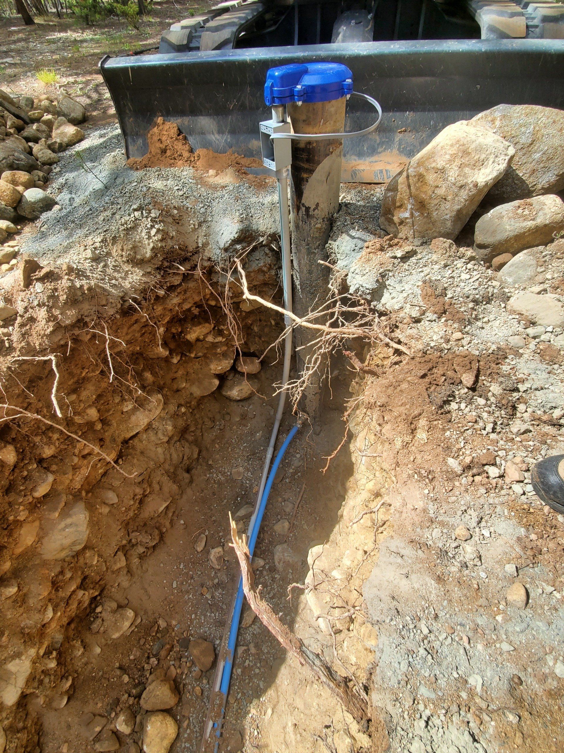 A trench in the ground with a water meter and pipes. Brown soil, rocks, and a tractor bucket are visible.