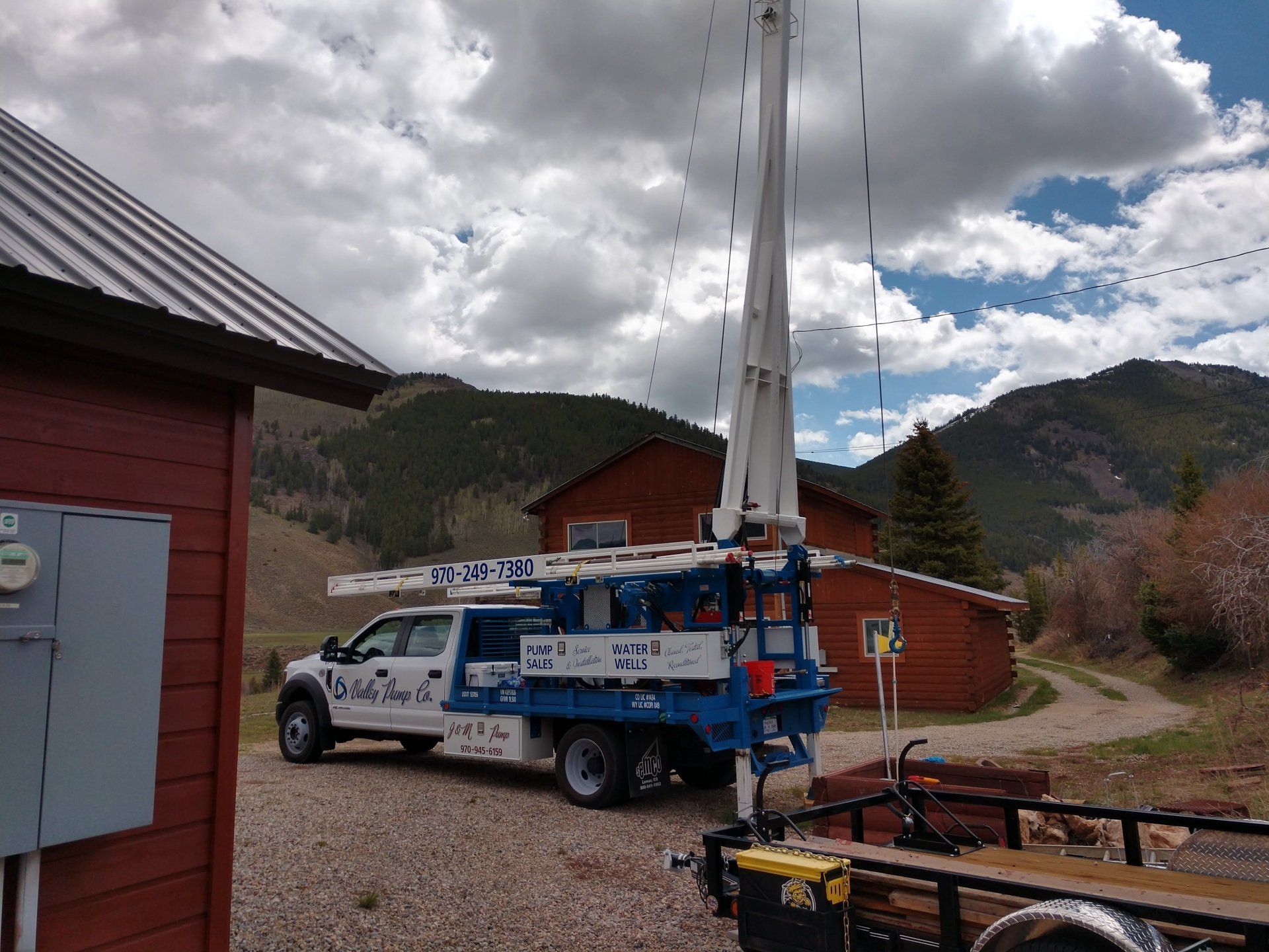 A white and blue drilling truck near a red building with a mountain background. The truck has a tall, white drill in the air.