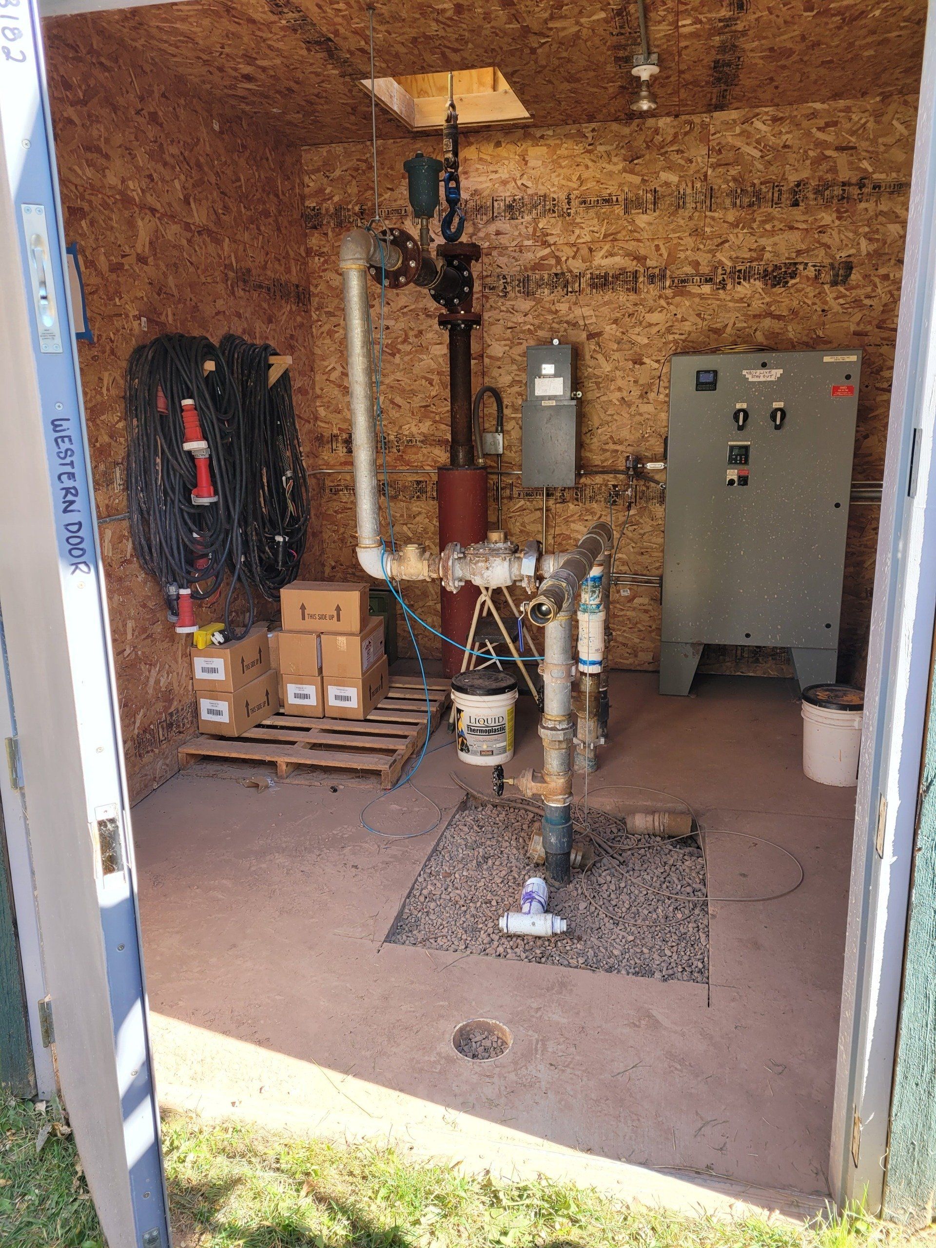 Interior of a small utility shed containing water pump equipment. Walls are wood, floor has gravel.