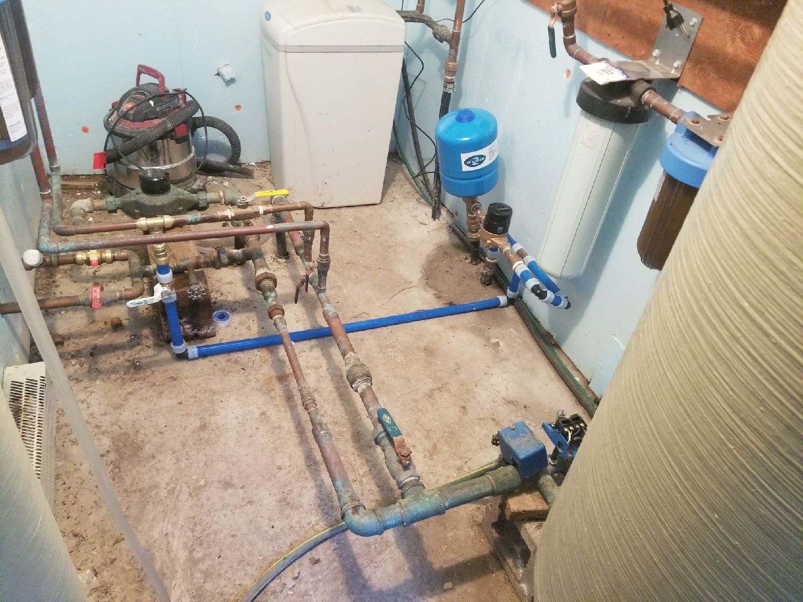 A water treatment system in a basement. Pipes, tanks, and a pump are visible. The setting appears functional with a concrete floor and blue walls.