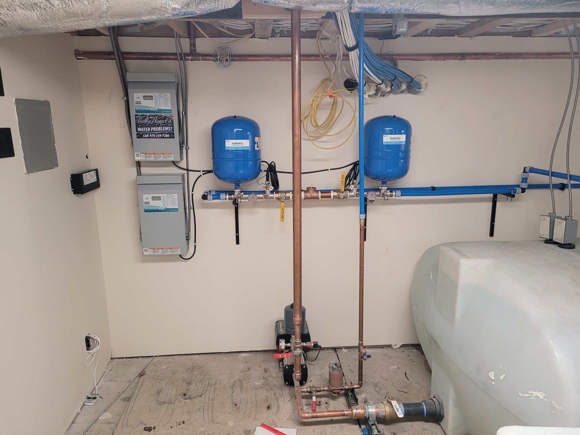 Plumbing system in a basement, featuring water tanks, copper pipes, pressure tanks, and control panels against a light-colored wall.