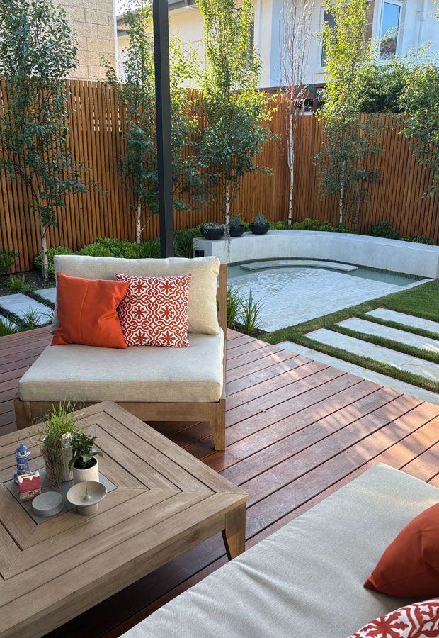 Poolside lounge area with white furniture under a wooden pergola, surrounded by lush greenery.