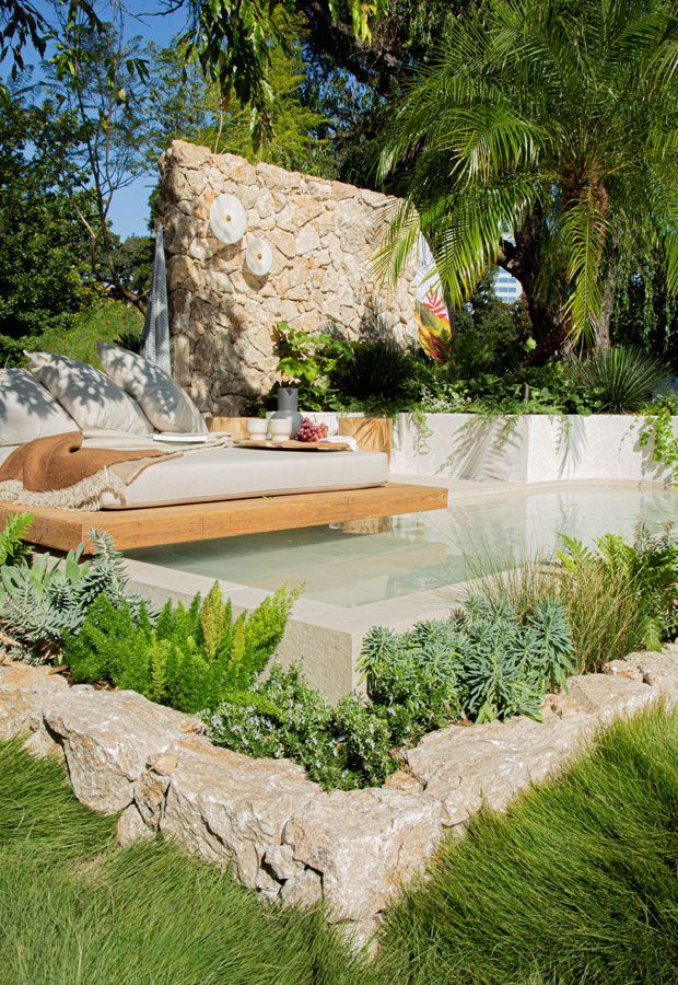 Modern house with pool, shaded patio, and wooden pergola. Turquoise water, glass walls, and dark wood siding.