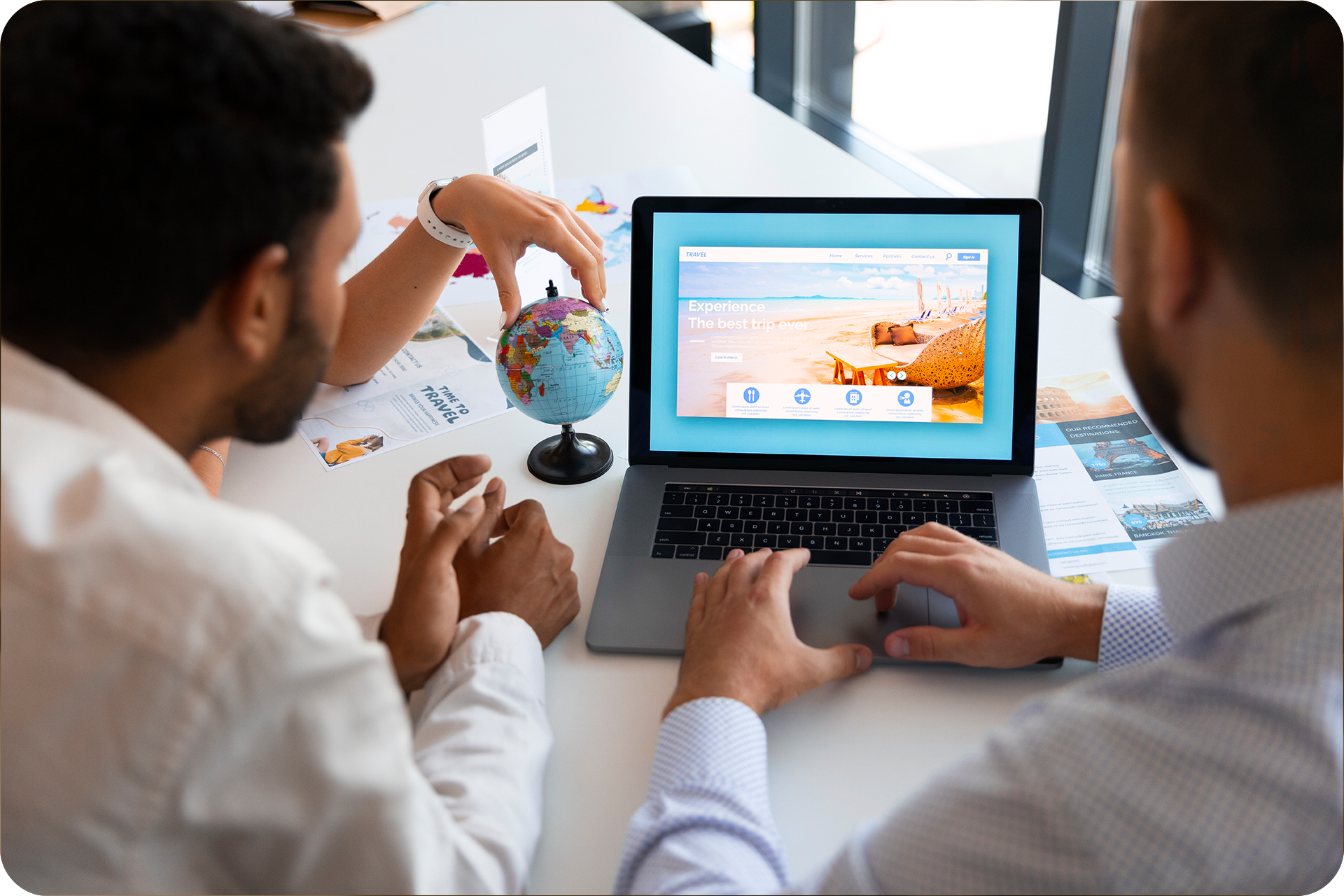 Duas pessoas em uma mesa olhando para um laptop exibindo uma cena de praia, com um globo ao fundo.