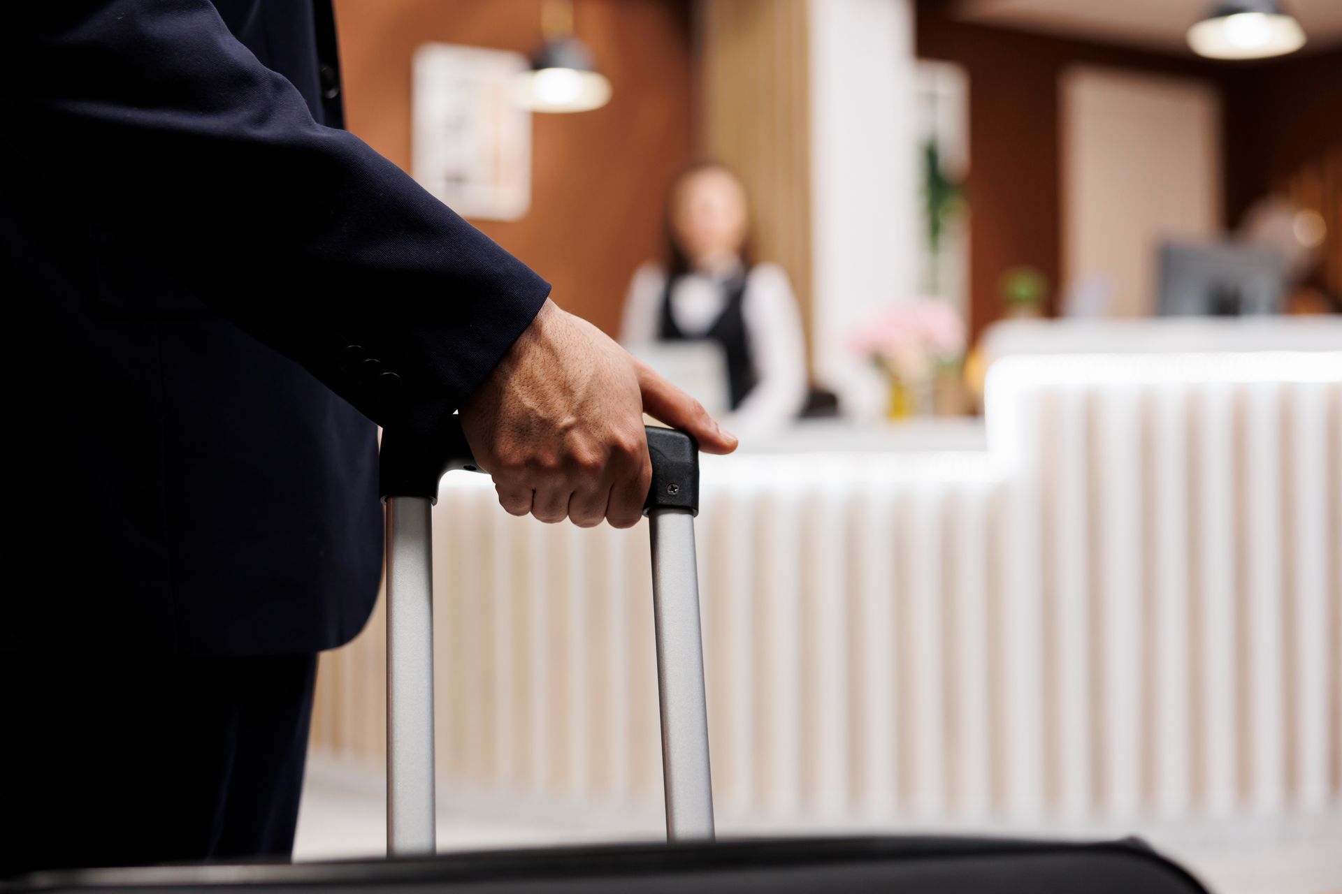 Homem de terno puxa uma mala em direção à recepção do hotel; um recepcionista está atrás.