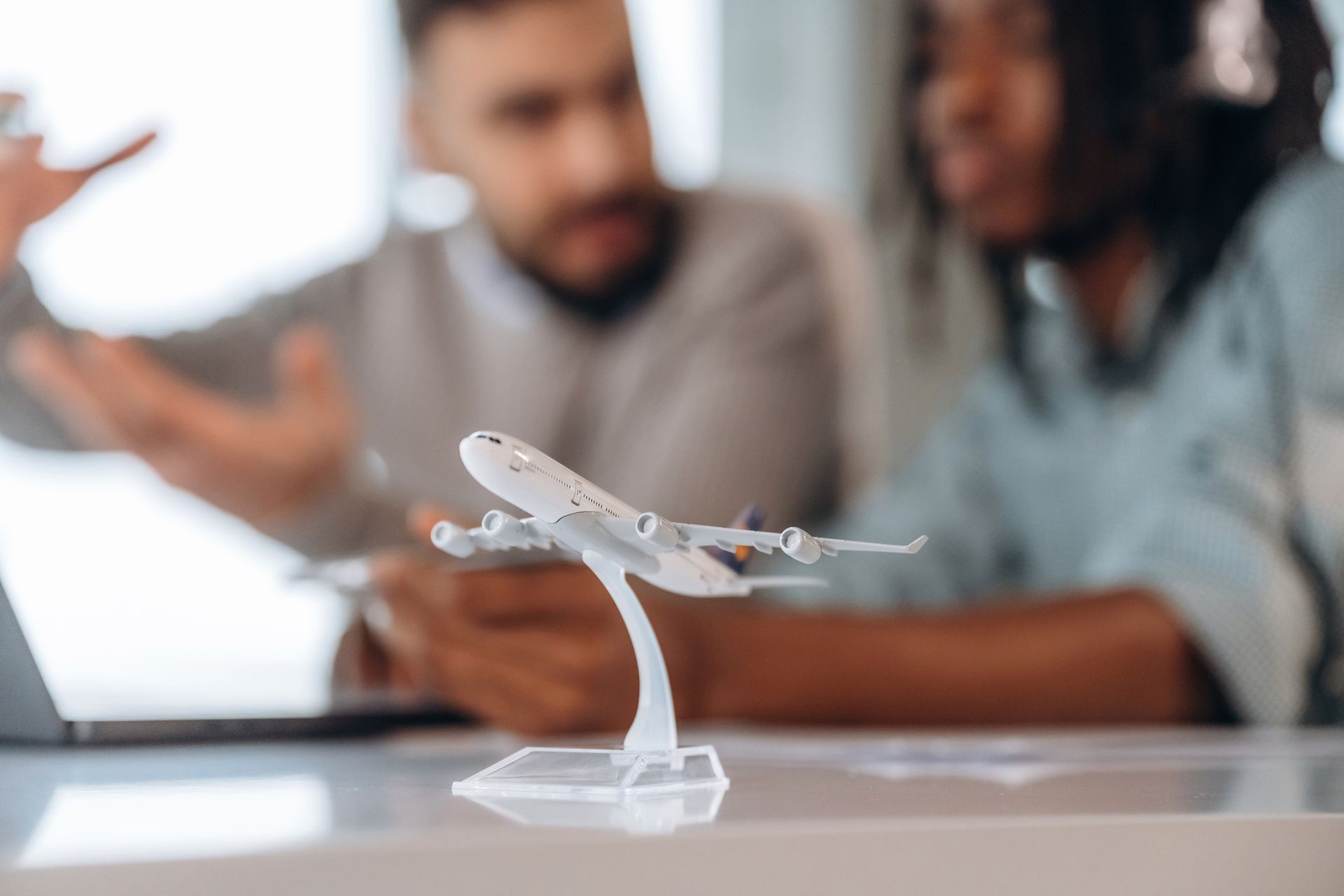 Homem gesticulando, mulher olhando para um laptop com um aeromodelo na mesa, reunião focada.