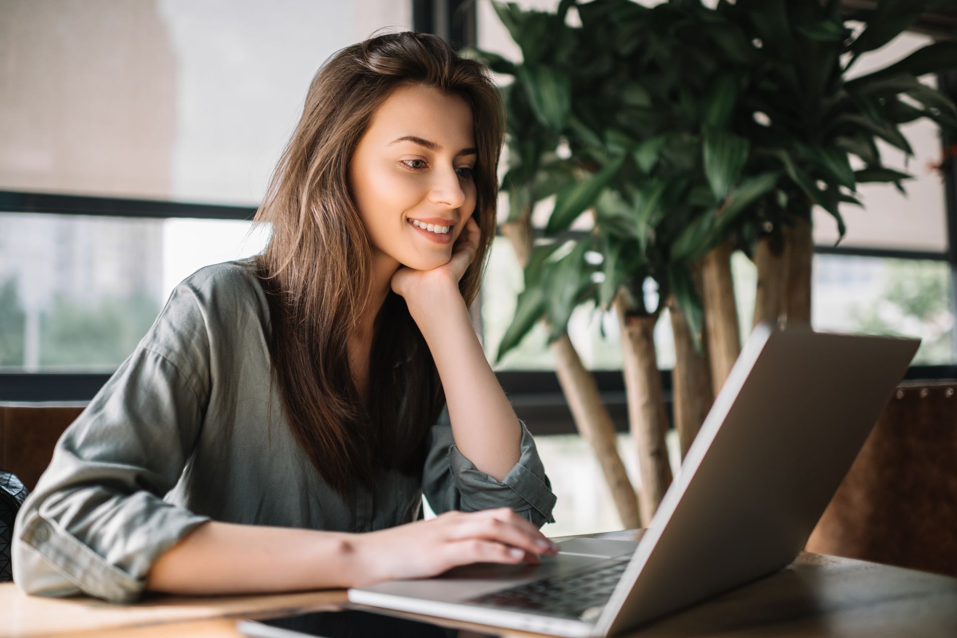Woman smiling at a laptop in a bright office, with a large plant in the background.