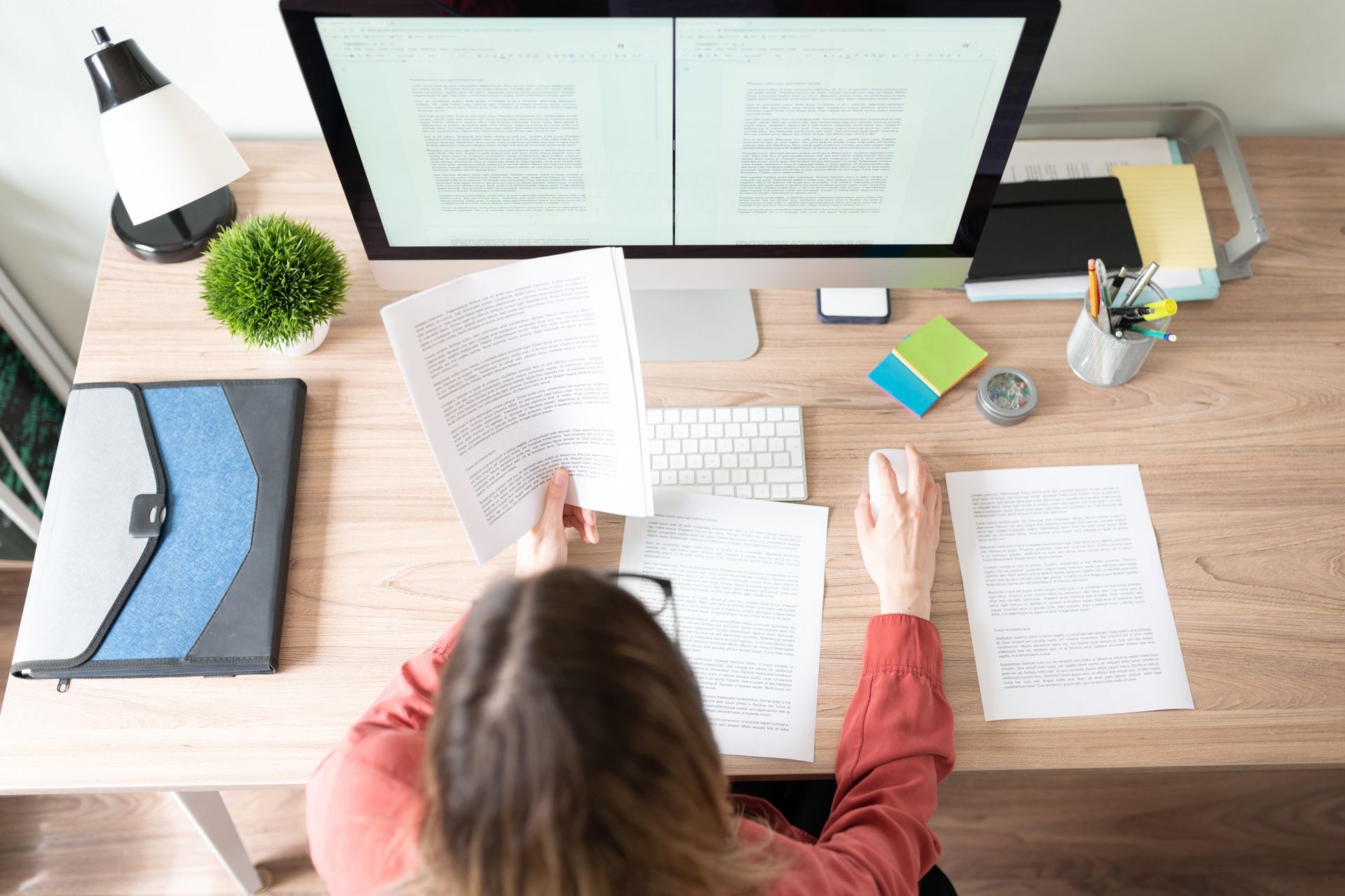 Overhead view of a desk with a person reviewing papers beside dual monitors, sticky notes, and office supplies