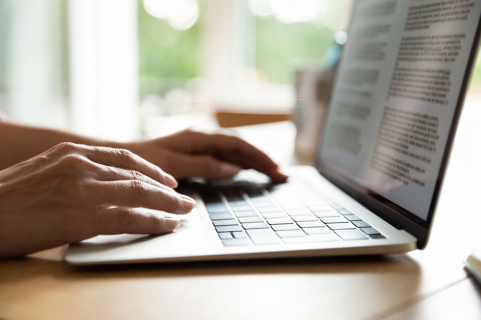 Hands typing on a laptop beside an open book on a wooden desk
