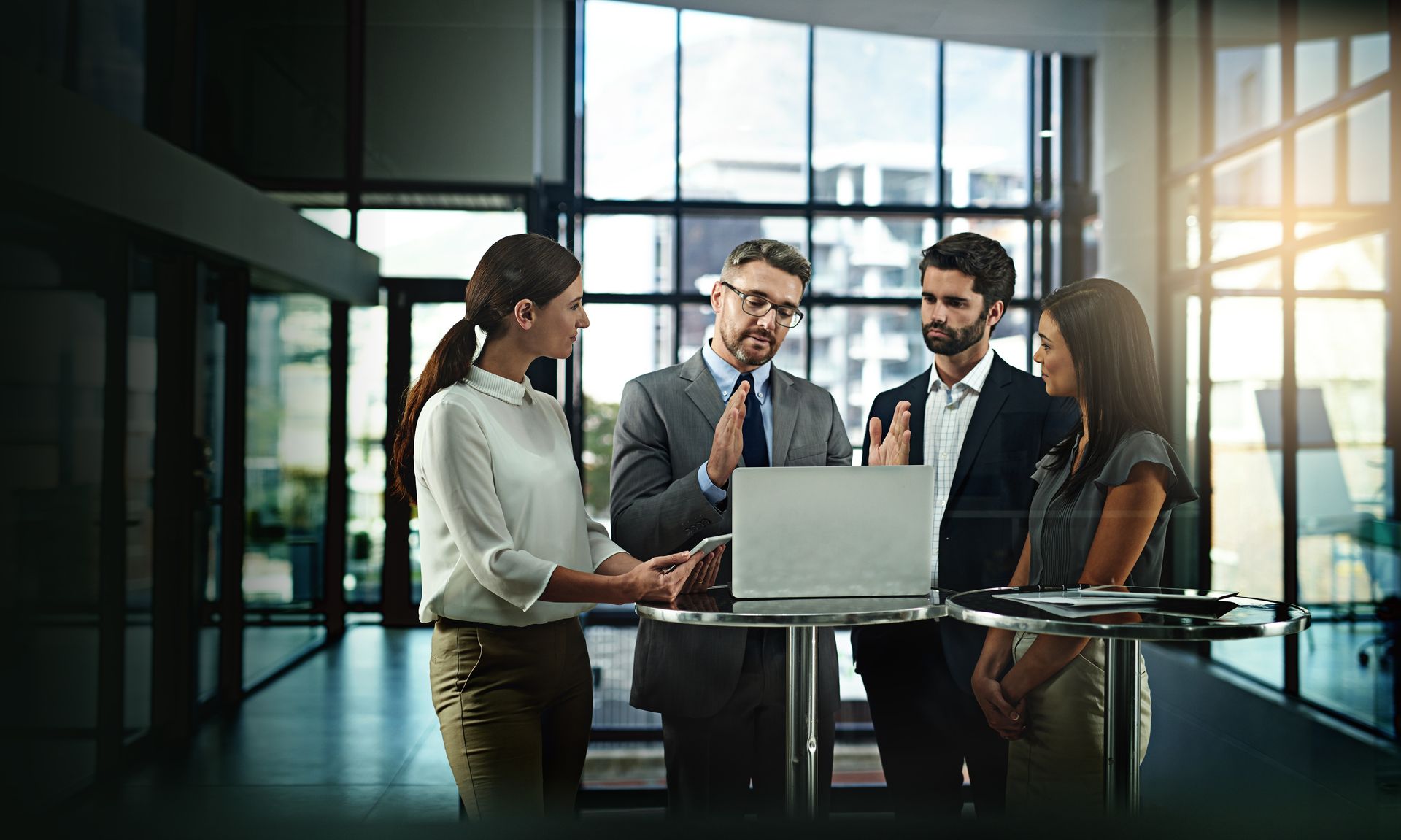 Four coworkers in business attire discuss around a laptop in a bright modern office.