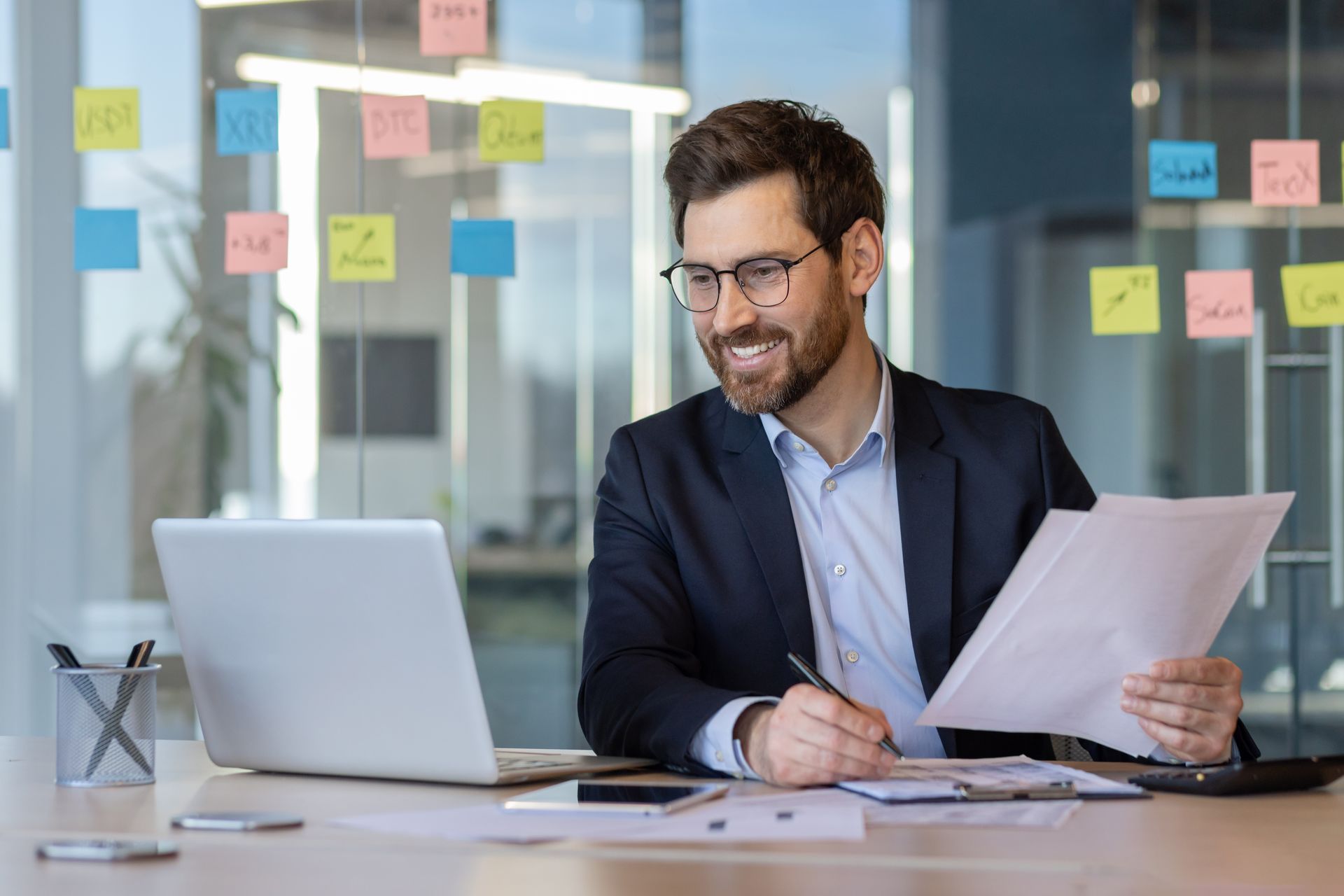 Man in a suit smiling at a desk with a laptop and papers in a glass-walled office with sticky notes on the wall