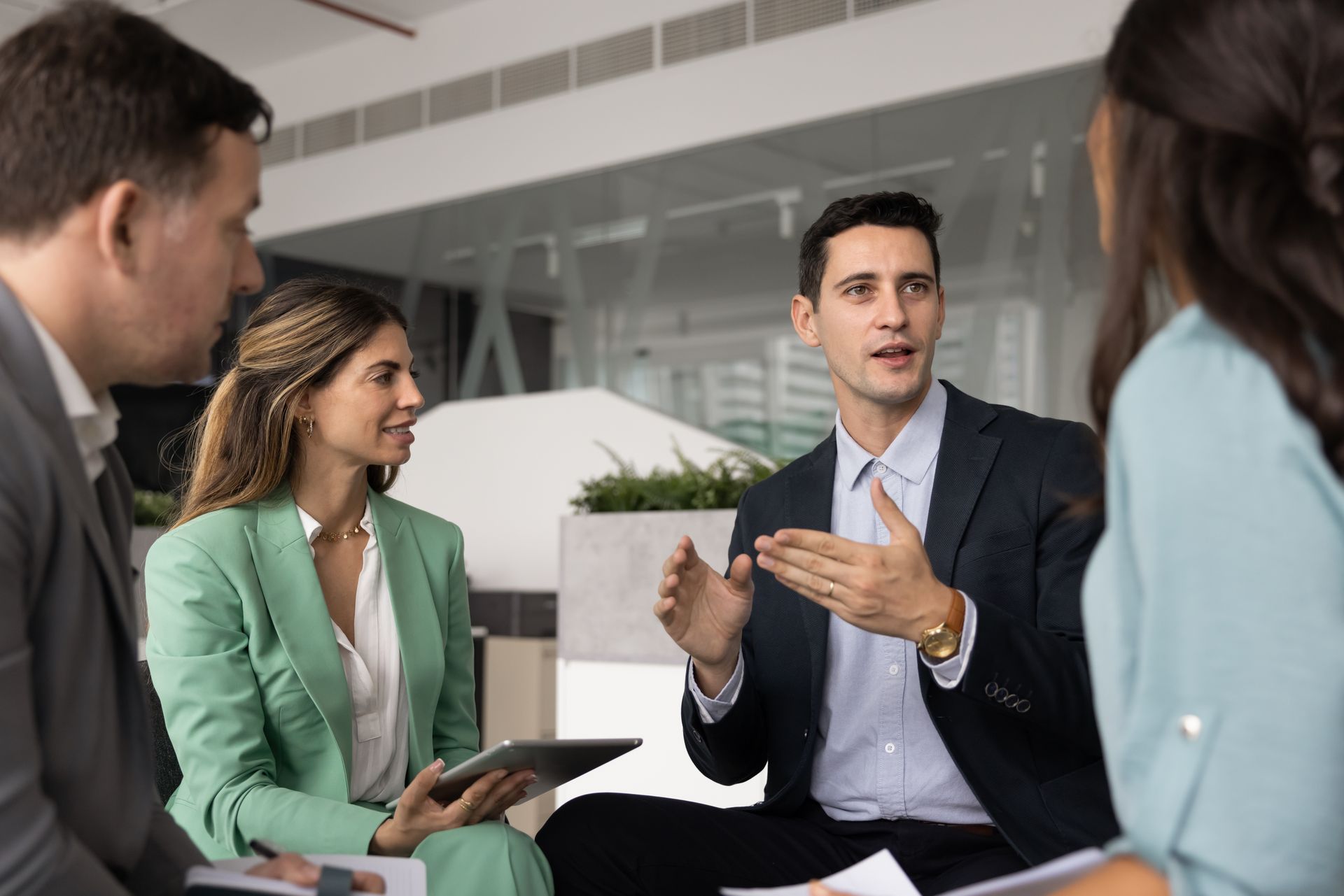 Business meeting with four people discussing around a table in a modern office, holding a tablet and gesturing.