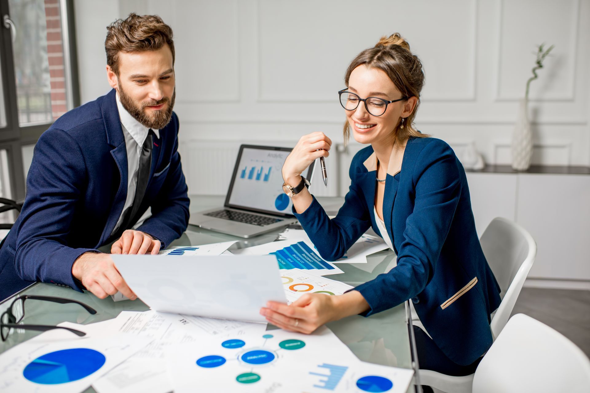 Two coworkers review charts at a desk with a laptop in a bright office
