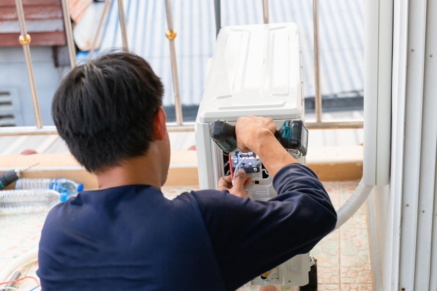 A technician in a navy shirt repairs the electrical wiring of an outdoor air conditioning unit on a balcony.