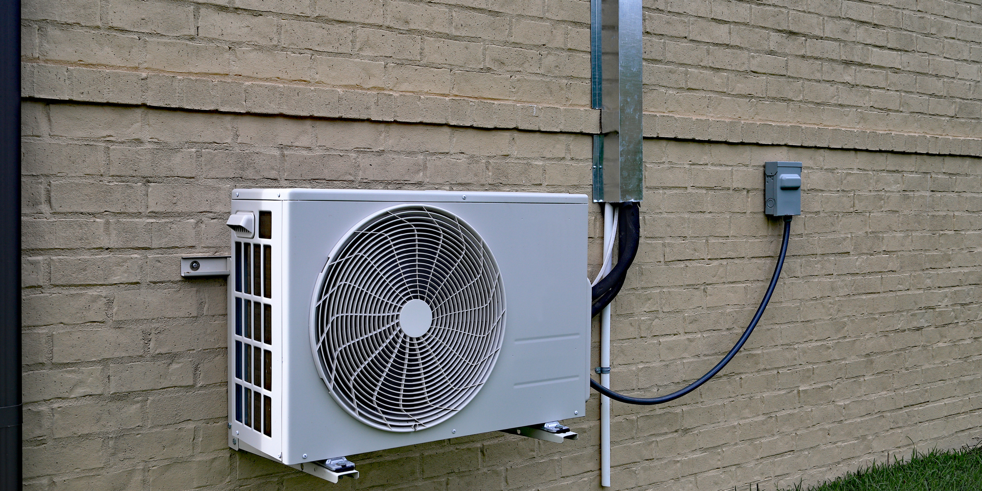 An outdoor HVAC unit mounted on a tan brick wall with an adjacent metal electrical conduit and disconnect box.