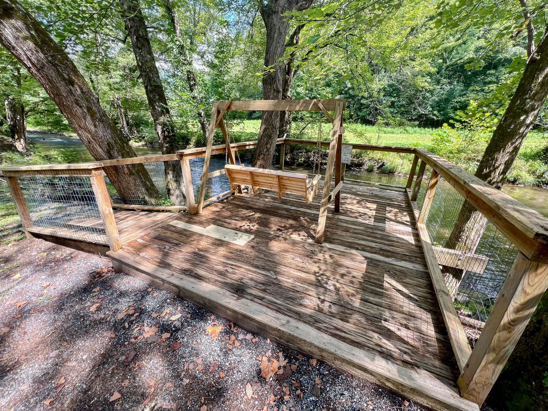 Wooden bridge over a creek, with a swing in the middle, surrounded by trees and greenery.