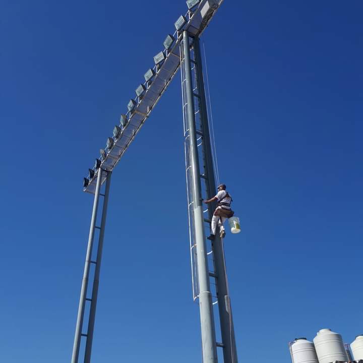 Persona subiendo a una alta torre de iluminación para realizar tareas de mantenimiento contra un cielo azul brillante.