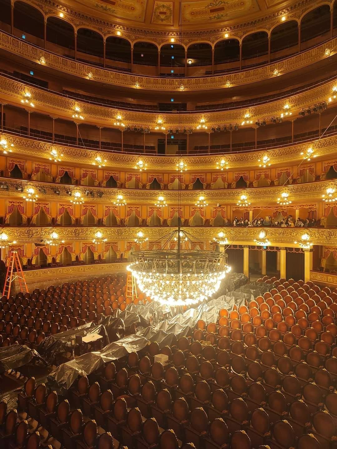 Interior de teatro ornamentado, filas de asientos vacíos, múltiples balcones, gran lámpara de araña.