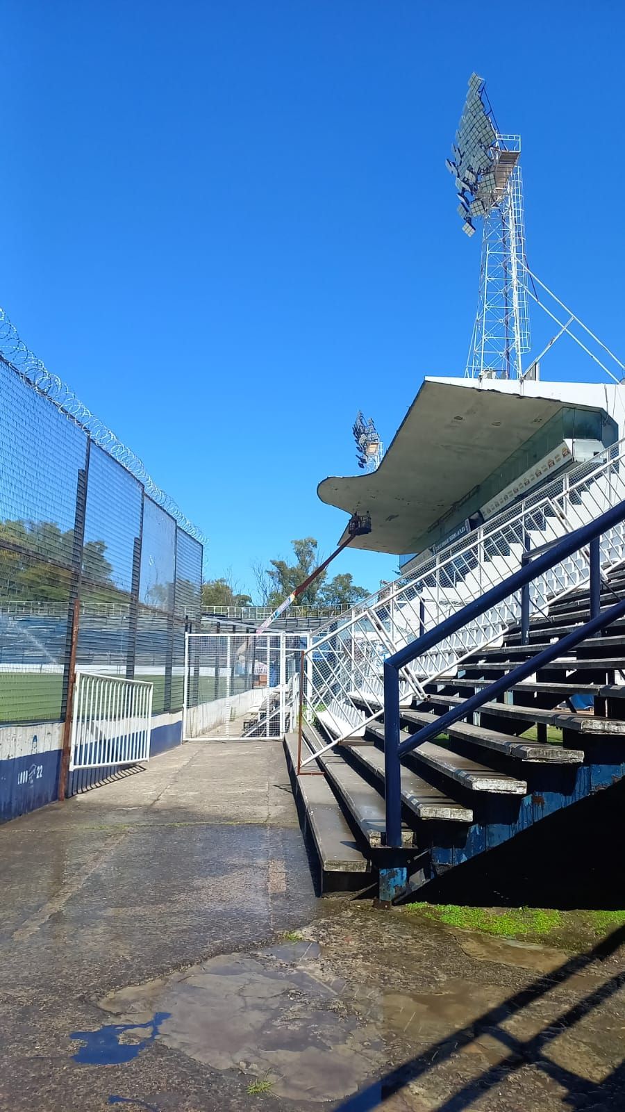 Gradas y pasarela del estadio en un día soleado, con una valla de alambre y una torre de iluminación.