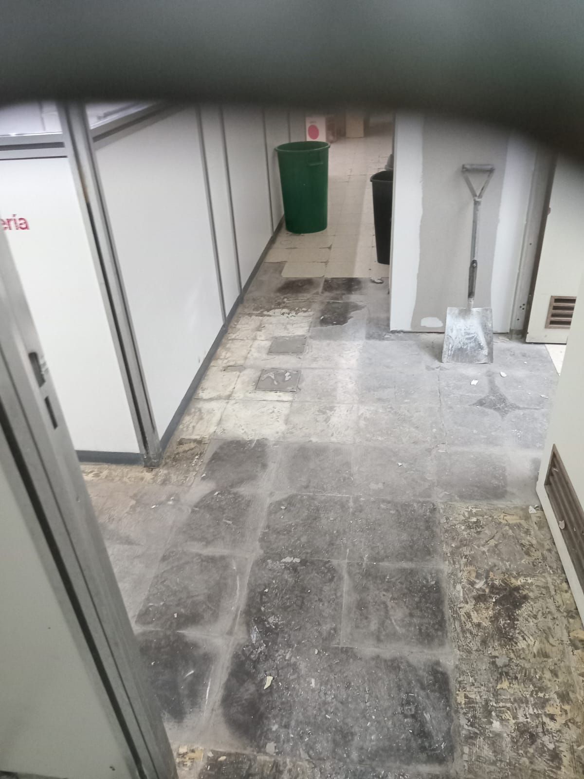 Un pasillo con baldosas grises dañadas, un bote de basura verde y una pala.