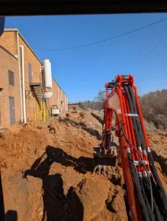 Excavator digging a trench beside a brick building on a dirt construction site under a clear blue sky Excavator digging a trench beside a brick building on a dirt construction site under a clear blue sky