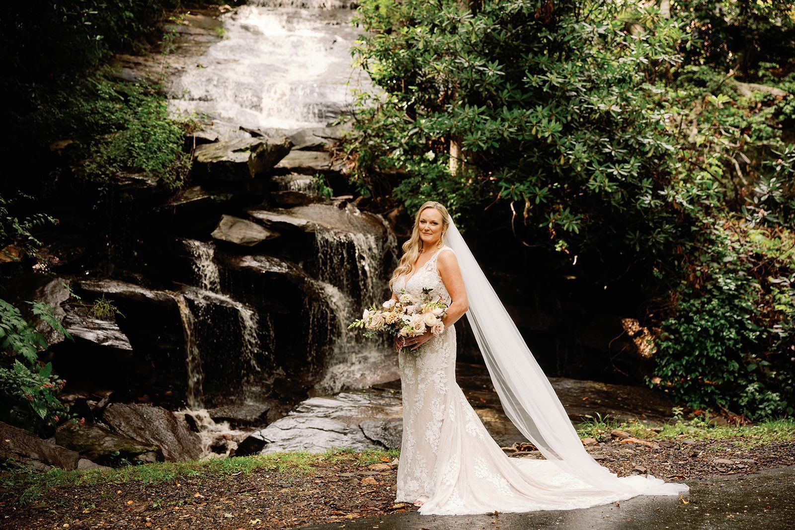 Bride in white gown holding flowers, posing by a small waterfall.