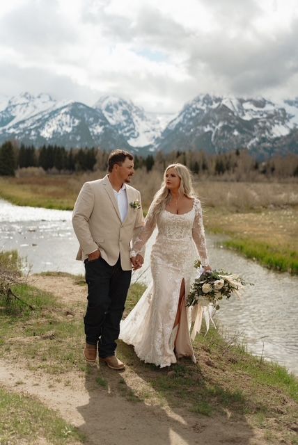 Couple in wedding attire hold hands by a stream, snow-capped mountains in the background.