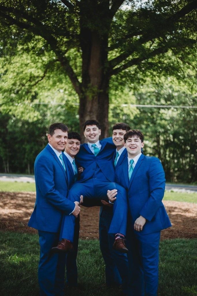 Five men in blue suits, smiling, holding one man up in front of a tree.