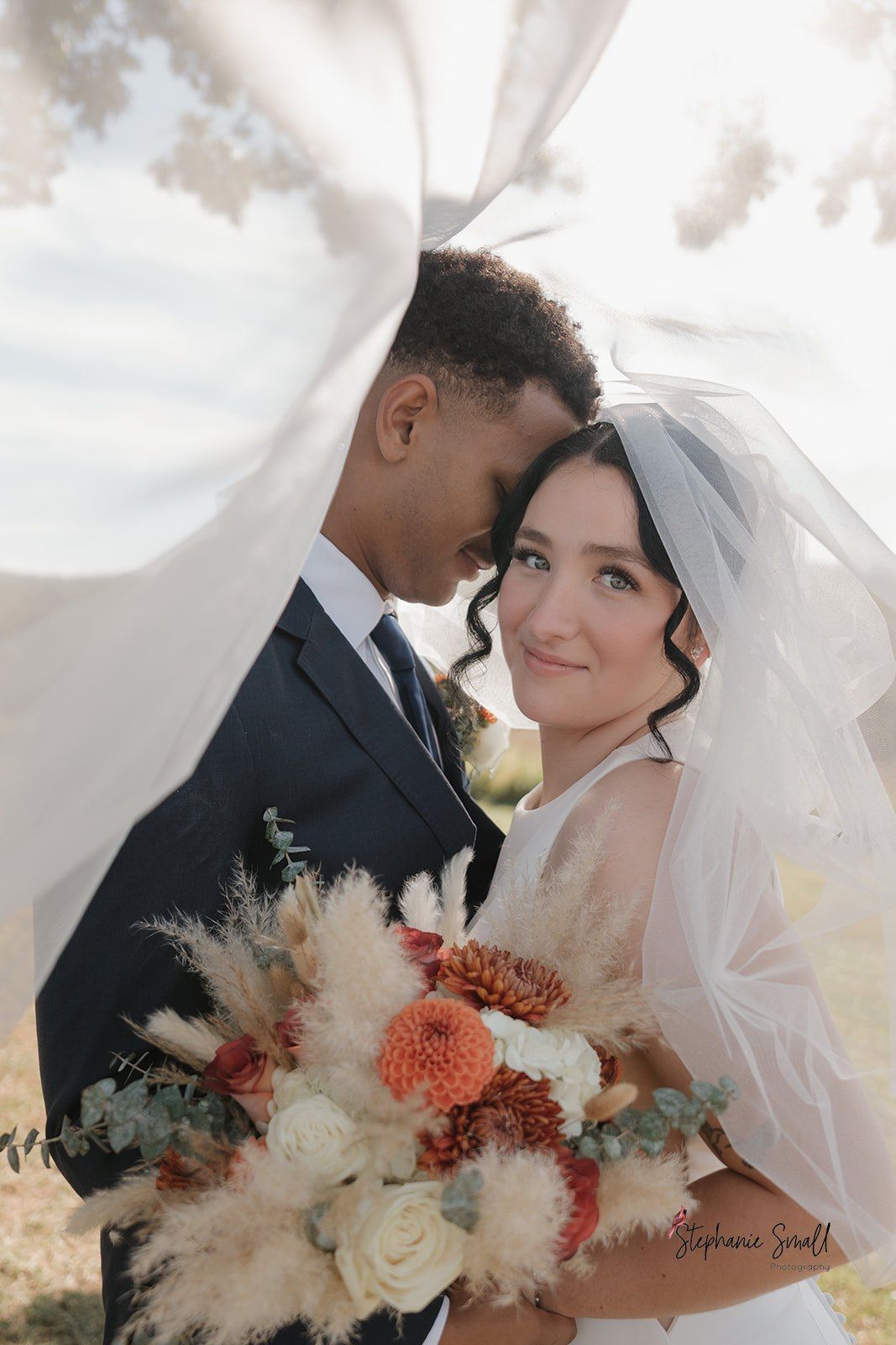 Bride and groom under a flowing veil, holding a bouquet. Outdoors.