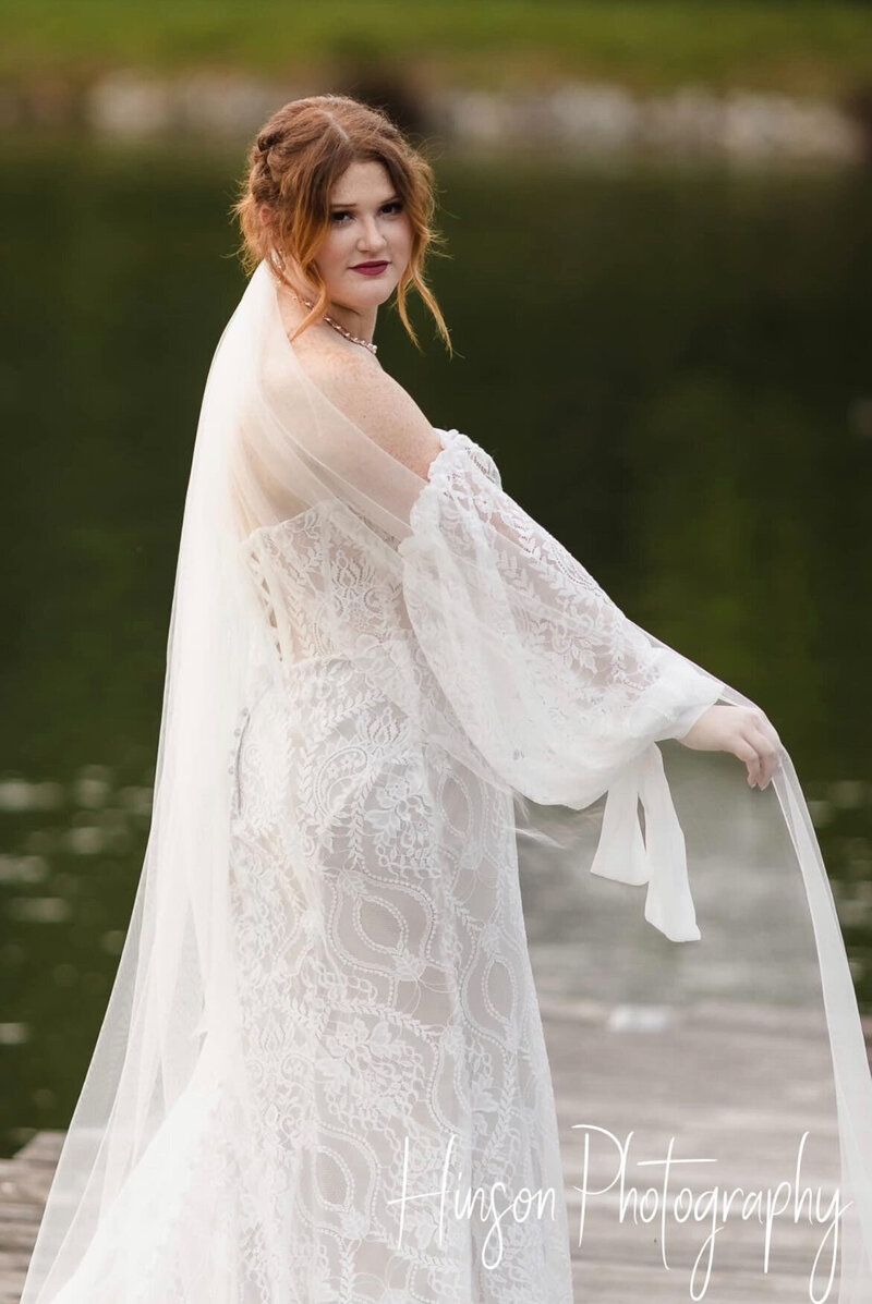 Woman in white lace wedding dress on wooden dock, holding veil, near water.