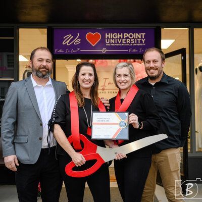Four people at a grand opening, holding giant scissors and a certificate, in front of a High Point University sign.