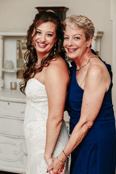 Bride in white dress holds hands with woman in blue dress, both smiling.