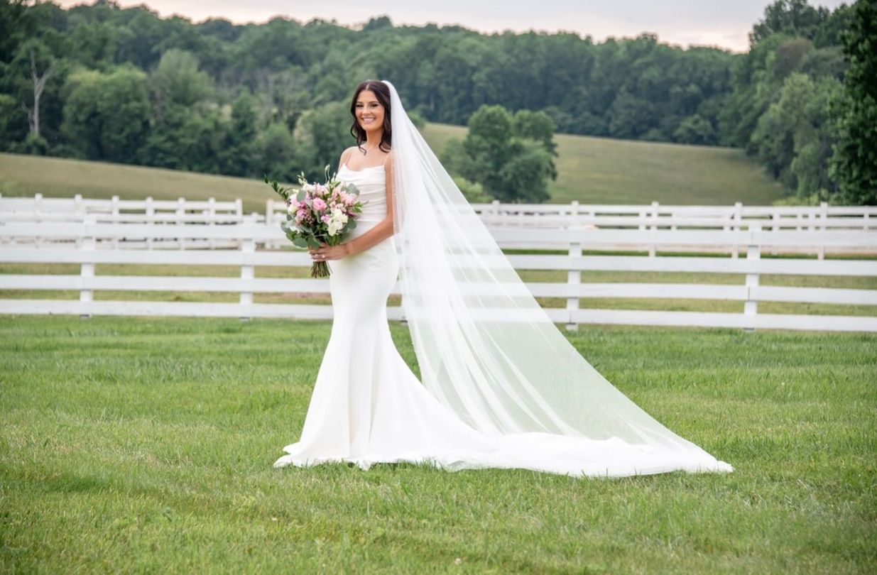Bride in a white gown and veil holds bouquet, standing on green grass, white fence and trees in background.